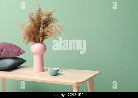 Hölzerner Couchtisch mit einer Tasse Tee, Vase mit Pampasgras und Kissen in der Nähe der grünen Wand im Zimmer Stockfoto