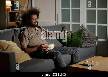 Junger birassischer Kerl, der entspannt auf der Couch im Wohnzimmer sitzt und Fernsehsender wechselt, Kopierraum Stockfoto