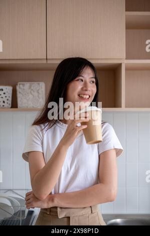 Eine schöne, positive junge asiatische Frau blickt auf die Aussicht nach draußen, während sie in der Küche Kaffee trinkt. Frühstück, Getränke, Lebensstil, Hausleben Stockfoto