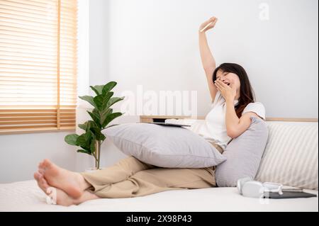 Eine attraktive junge asiatische Frau, die bequeme Kleidung trägt, gähnt und dehnt ihre Hände, nachdem sie ihr digitales Tablet benutzt hat, während sie in einer Co-Wohnung sitzt Stockfoto