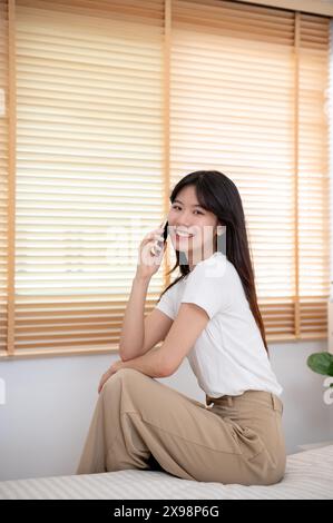 Eine attraktive junge asiatische Frau telefoniert mit jemandem, während sie im Schlafzimmer auf ihrem Bett sitzt. Konzepte für Menschen und drahtlose Technologie Stockfoto