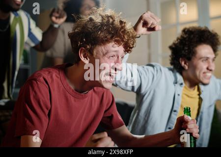 Selektive Fokusaufnahme eines fröhlichen jungen Kaukasiers und seiner multiethnischen Freunde, die Sportveranstaltungen im Fernsehen verfolgen Stockfoto