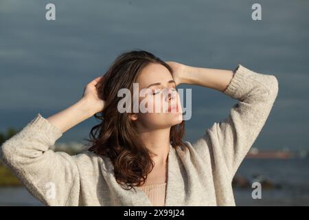 Genießen Sie die brünette Frau mit frischer Haut, natürlichem Make-up und gesundem, welligen braunen Haar, das die sonnige Morgen- und Meeresluft genießt. Weibliches Modell für den Außenbereich Stockfoto