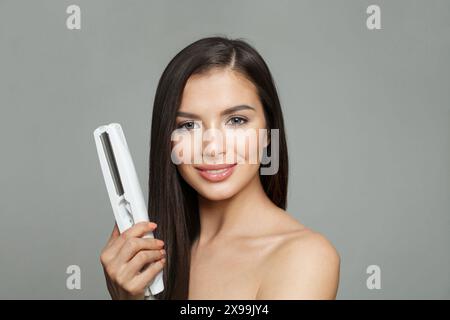 Haarglätterkonzept. Junge gesunde Brünette Frau, die Haar Eisen hält und ihr langes glattes glänzendes Haar auf weißem Hintergrund glättet Stockfoto