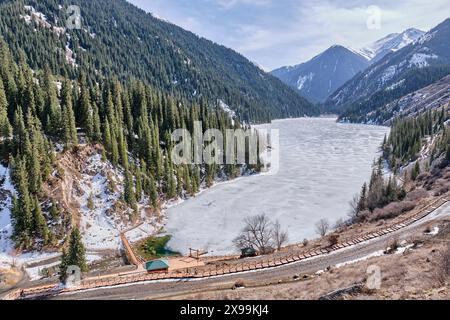 Almaty, Kasachstan - 19. März 2024: Malerischer Kolsai-See mit Eis bedeckt, umgeben von grünen Hügeln. Fußgängerzone mit hölzernem Öko-Pfad rund um den See. Feder Stockfoto