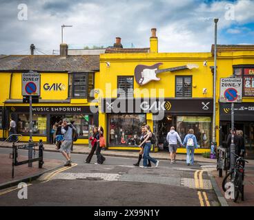 Die Leute laufen vorbei am gelb bemalten Gak Music Emporium und dem Gitarrenladen im Lanes Conservation Area in Brighton, East Sussex, Großbritannien. Stockfoto