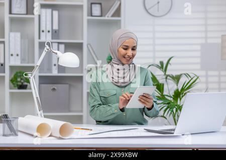 Selbstbewusste Architektin, die Hijab trägt und an ihrem Schreibtisch in einer modernen Büroumgebung auf einem Tablet arbeitet. Professionell und konzentriert sich auf ihre Architekturprojekte. Stockfoto