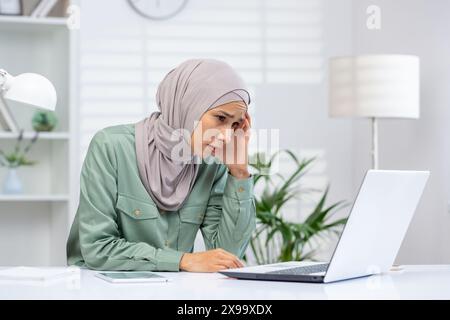 Gestresste Geschäftsfrau im Hijab, die in einer modernen Büroumgebung an einem Laptop arbeitet. Konzept von Stress am Arbeitsplatz, Herausforderungen und Druck im beruflichen Umfeld. Stockfoto