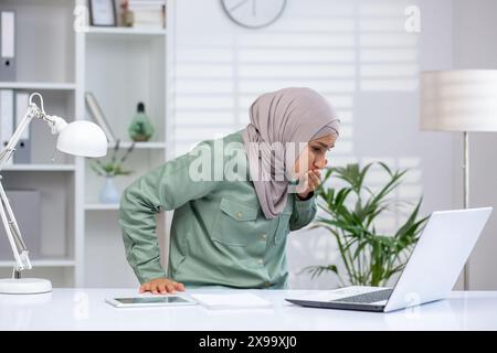 Eine Frau in einem Hijab, die sich krank fühlt und Übelkeit erleidet, während sie an ihrem Schreibtisch in einem Büro arbeitet und ihren Laptop anstarrt. Stockfoto