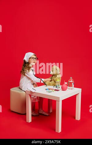 Als Arzt gekleidetes Kind, das mit medizinischen Vorräten am Tisch sitzt, untersucht Teddybär-Patienten vor leuchtendem rotem Hintergrund. Stockfoto