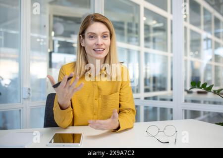 Professionelle Frau, die in einer modernen Büroumgebung Videoanrufe führt und während des Sprechens direkt auf die Kamera blickt. Stockfoto