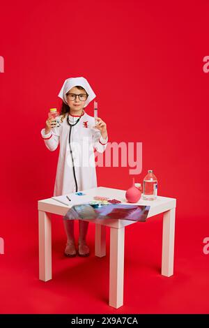 Kleiner Arzt. Kind in medizinischer Kleidung gekleidet, posiert neben Tisch mit verschiedenen medizinischen Vorräten vor lebendigem rotem Hintergrund. Stockfoto