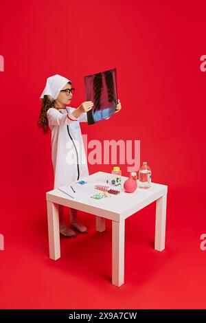 Mädchen, das als Arzt gekleidet ist, untersucht Röntgenaufnahmen, stehend am Tisch mit medizinischen Vorräten vor leuchtend rotem Hintergrund. Stockfoto