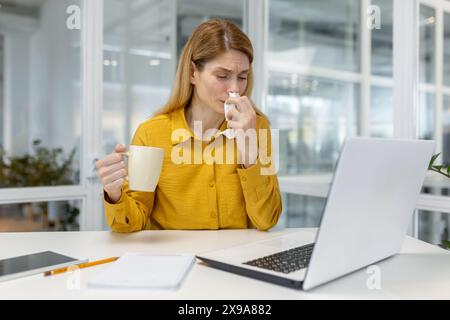 Eine Frau in einem gelben Hemd sitzt an ihrem Schreibtisch, trinkt Tee und bläst sich die Nase, was auf Krankheit und Unwohlsein bei der Arbeit hinweist. Stockfoto