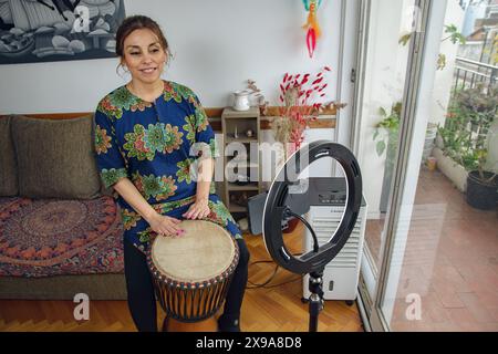 Erwachsene lateinamerikanische Frau genießt den Morgen mit ihrer Djembe-Trommel, macht Musik und filmt sich mit einem Mobiltelefon. Lächelt glücklich und sie ist Attenti Stockfoto