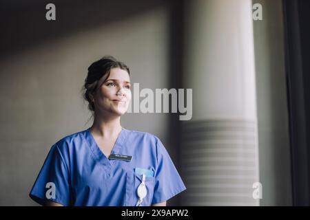 Lächelnde nachdenkliche Ärztin in blauen Schutten im Krankenhaus Stockfoto