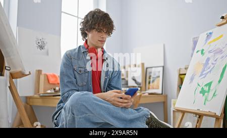 Ein junger Mann mit lockigen Haaren und Kopfhörern benutzt ein Smartphone in einem hellen Kunststudio. Stockfoto