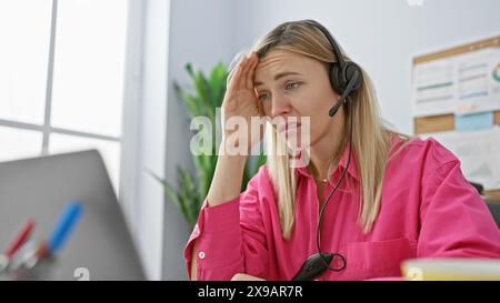 Eine gestresste junge blonde Frau, die ein Headset in einem hellen Büro trägt und Frustration und Erschöpfung am Arbeitsplatz darstellt. Stockfoto