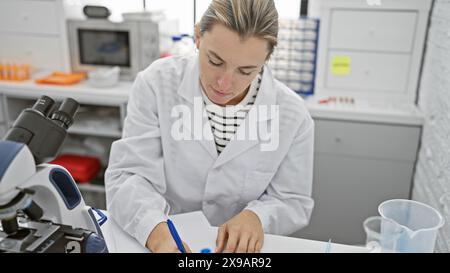 Eine fokussierte junge Wissenschaftlerin schreibt Notizen im Labor mit Mikroskop und Laborausrüstung. Stockfoto