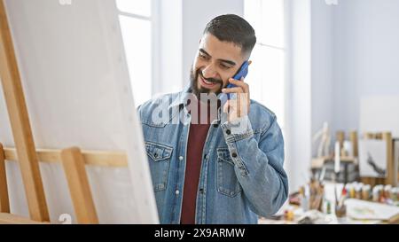 Lächelnder bärtiger Mann in Jeansjacke, der im Kunststudio telefoniert, umgeben von Gemälden und Kunstbedarf Stockfoto