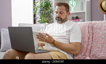 Ein hispanischer Mann mittleren Alters mit grauen Haaren, mit Kopfhörern, schreibt in einem Notizbuch auf einem Sofa drinnen. Stockfoto