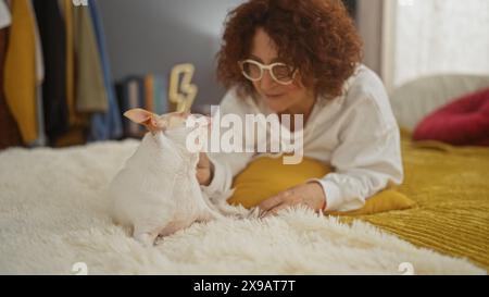 Eine Frau mittleren Alters mit lockigen Haaren liegt auf einem Bett in einem gemütlichen Schlafzimmer und streichelt liebevoll ihren chihuahua, umgeben von warmen Innenräumen. Stockfoto