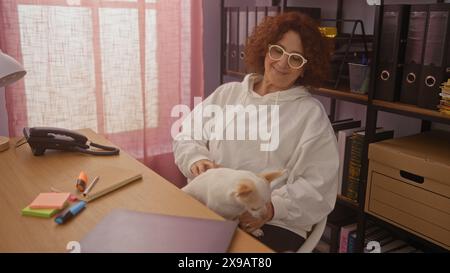 Frau mittleren Alters in einem Heimbüro, lächelt mit ihrem chihuahua-Hund auf dem Schoß, umgeben von Bürobedarf und Regalen mit Akten. Stockfoto