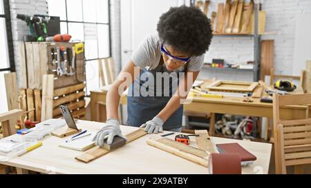 Fokussierte schwarze Frau, die Holz in einer gut ausgestatteten Tischlerei schleift, mit Schutzbrille. Stockfoto