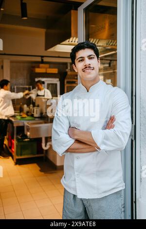 Porträt eines glücklichen männlichen Küchenchefs mit überkreuzten Armen, der in der Geschäftsküche an der Tür steht Stockfoto