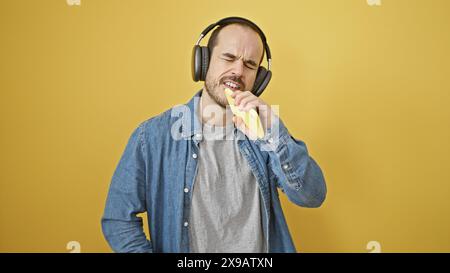 Ein bärtiger kahlköpfiger Mann singt in ein Telefon mit Kopfhörern vor gelbem Hintergrund. Stockfoto