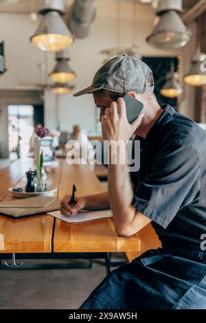 Männlicher Besitzer schreibt auf Notizblock, während er im Café mit dem Smartphone telefoniert Stockfoto