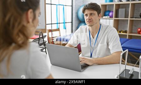 Ein Arzt und eine Patientin in einer Reha-Klinik diskutieren über die Behandlung über einen Laptop in einem hellen Therapieraum. Stockfoto