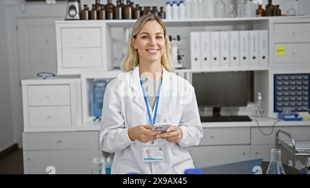 Selbstbewusste junge kaukasische Wissenschaftlerin mit blonden Haaren im weißen Labormantel mit Smartphone im Labor. Stockfoto