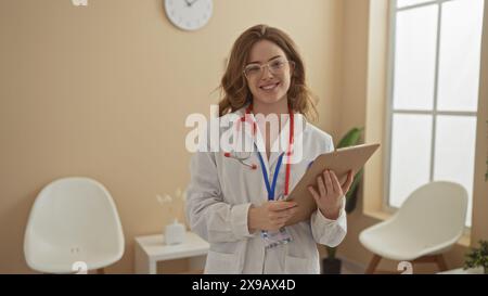 Eine schöne junge blonde Ärztin steht selbstbewusst in einem Wartezimmer des Krankenhauses, hält ein Klemmbrett und lächelt warm. Stockfoto