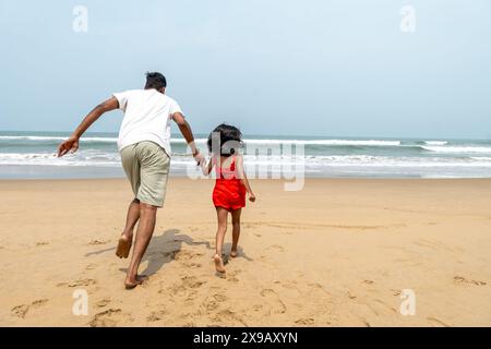 Ein indischer Vater und seine kleine Tochter sprinten fröhlich über den Sandstrand, ihr Lachen ertönt, während sie auf das sprudelnde Wasser zurennen. Stockfoto