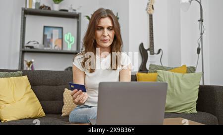 Reife Frau mit Kreditkarte, während sie im modernen Wohnzimmer einen Laptop benutzte, elektronisches Einkaufskonzept. Stockfoto