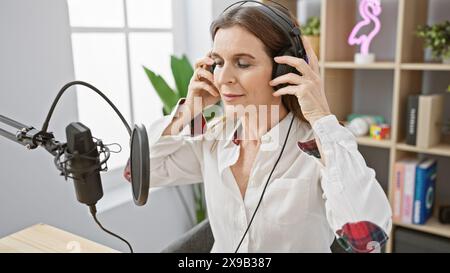 Die reife Frau nimmt Podcasts im Studio auf und trägt Kopfhörer neben Mikrofon und Tontechnik Stockfoto