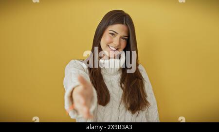 Eine fröhliche junge hispanische Frau in einem weißen Pullover, die einen Handschlag vor gelbem Hintergrund anbietet. Stockfoto