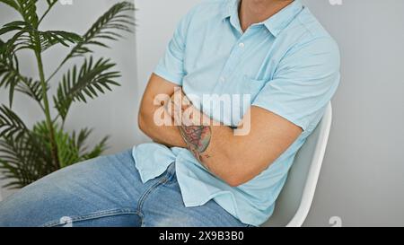 Ein junger hispanischer Mann mit Bart und Tätowierung sitzt in einem hellblauen Hemd und einer Jeans drinnen im Kreuz. Stockfoto