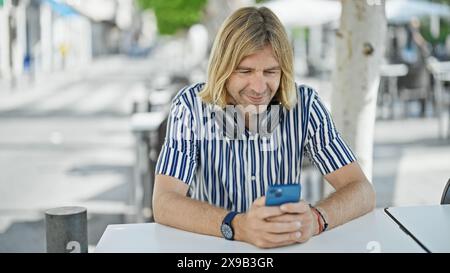 Ein blonder Mann mit Kopfhörern lächelt, als er sein Smartphone in einem Café im Freien in einer europäischen Stadt benutzt. Stockfoto