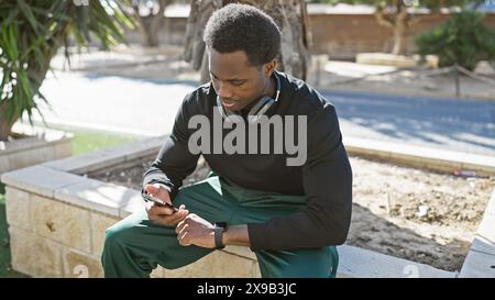 Ein junger afroamerikaner mit Kopfhörern und Smartwatch sucht sein Handy in einem sonnigen Park. Stockfoto