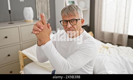 Ein reifer, grauhaariger Mann macht eine Handfeuergeste in einem Schlafzimmer und vermittelt Verspieltheit oder metaphorische Konzepte. Stockfoto