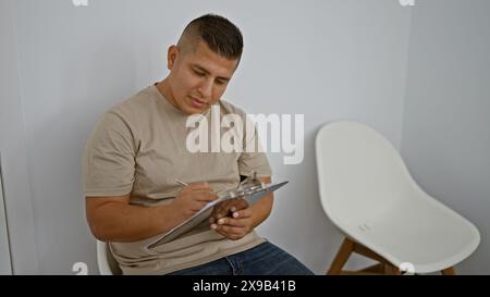 Ernsthafter lateinmann, entspannt und konzentriert, sitzt im Stuhl und macht Notizen. Innenporträt eines jungen hispanischen Mannes, der eine Checkliste für das Bewerbungsgespräch auf Clip schreibt Stockfoto