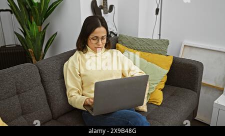 Hispanische Frau in Brille mit Laptop auf Couch im Wohnzimmer Stockfoto