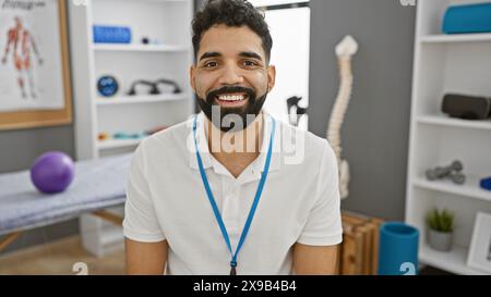 Ein hübscher junger Mann mit Bart lächelt herzlich, trägt ein Schlüsselband in einer gut ausgestatteten Physiotherapie-Klinik. Stockfoto