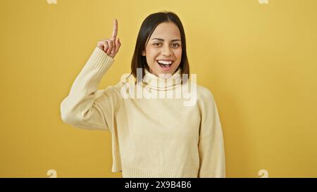 Fröhliche junge hispanische Frau in einem beigefarbenen Pullover, der vor einem leuchtend gelben Hintergrund zeigt. Stockfoto