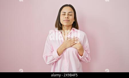 Ruhige hispanische Frau in rosa Hemd mit geschlossenen Augen auf einem glatten rosa Hintergrund, die Ruhe und Achtsamkeit vermittelt. Stockfoto