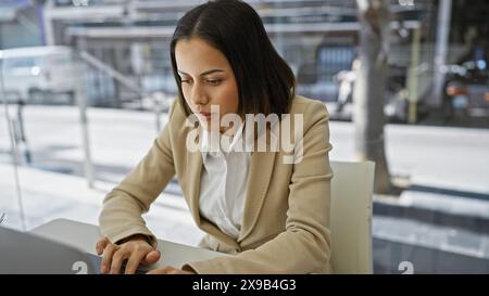 Eine fokussierte junge hispanische Geschäftsfrau arbeitet fleißig an ihrem Laptop in einer modernen Büroumgebung. Stockfoto