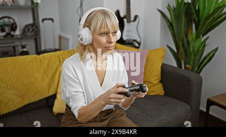 Konzentrierte blonde Frau beim Gaming mit Kopfhörer und Controller in einem gemütlichen Wohnzimmer. Stockfoto