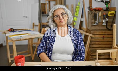 Reife Frau mit Brille in der Holzwerkstatt, umgeben von Werkzeug- und Möbelprojekten. Stockfoto